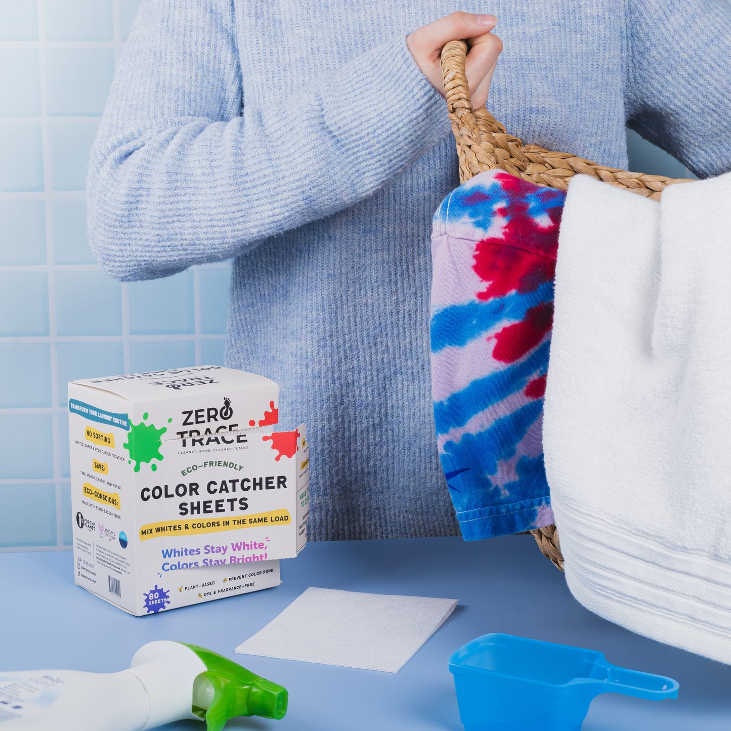 A person holds a laundry basket near a box of Zero Trace Color & Dye Protector Sheets, laundry detergent, and a spray bottle on a blue countertop—ideal for preventing color runs and keeping clothes vibrant.