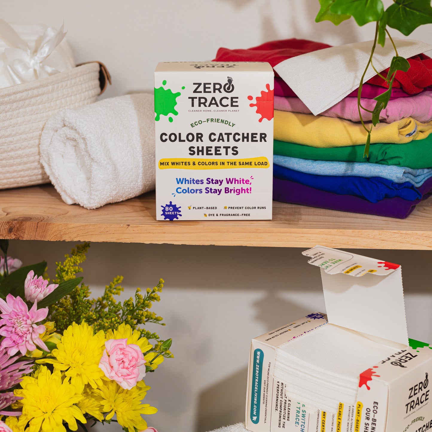 A box of Zero Trace Color & Dye Protector Sheets rests on a wooden shelf beside folded towels, stacked colorful clothes, and a bouquet of yellow and pink flowers, ensuring color run prevention with every wash.