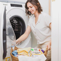 A woman with a laundry basket and a box of Zero Trace Color & Dye Protector Sheets opens her front-loading washing machine, ready to protect her clothes from unwanted dye transfer.