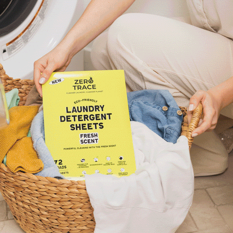 A person places a box of Zero Trace Laundry Detergent Sheets into a woven basket filled with clothes near the washing machine.