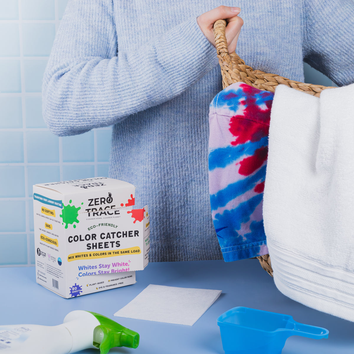 A person holds a laundry basket near a box of Zero Trace Color & Dye Protector Sheets, laundry detergent, and a spray bottle on a blue countertop—ideal for preventing color runs and keeping clothes vibrant.