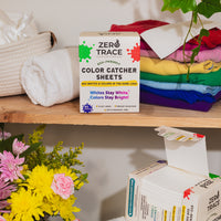 A box of Zero Trace Color & Dye Protector Sheets rests on a wooden shelf beside folded towels, stacked colorful clothes, and a bouquet of yellow and pink flowers, ensuring color run prevention with every wash.